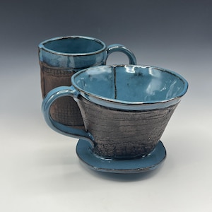 May include: Two handmade ceramic coffee cups. One is a standard mug, the other a pour-over coffee maker. Both have a textured dark gray exterior and a glossy teal blue interior and rim. The mug has a matching teal blue handle.