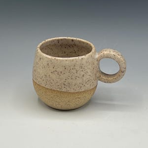 May include: A speckled ceramic mug with a rounded body and a circular handle. The top half of the mug is a light beige colour with brown speckles, while the bottom half is a lighter tan colour. The mug is designed for hot beverages.