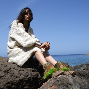 May include: A woman wearing a white knitted sweater sits on a rocky shore, looking out at the ocean. She is wearing green and brown boots with feathers.