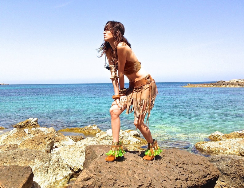 May include: A woman wearing a brown leather fringe skirt and a brown leather top with a gold chain. She is standing on a rocky beach with the ocean in the background.