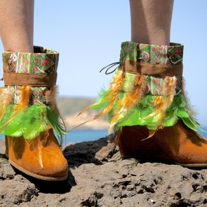 May include: Brown suede ankle boots with green and orange feather trim. The boots have a patterned fabric cuff and are worn on a rocky surface.