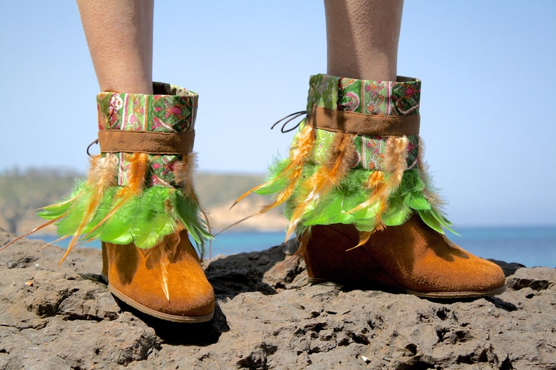 May include: Brown suede ankle boots with green and orange feather trim. The boots have a patterned fabric cuff and are worn on a rocky surface.