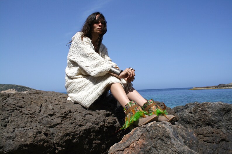 May include: A woman wearing a white knitted sweater sits on a rocky shore, looking out at the ocean. She is wearing green and brown boots with feathers.