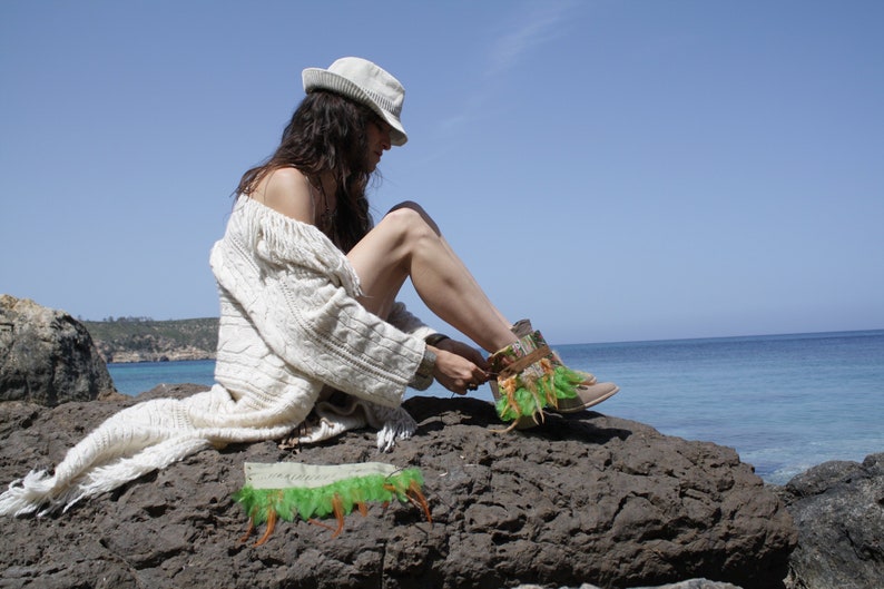 May include: A woman wearing a white sweater and a white hat sits on a rocky shore, tying the laces of a brown and green ankle boot with feathers.