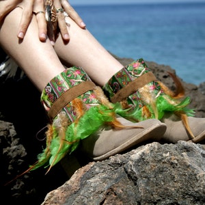 May include: A pair of brown suede ankle boots with green and orange feather trim. The boots are decorated with a colorful patterned fabric around the ankle.