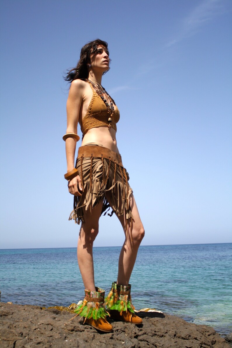 May include: A woman wearing a brown leather fringe skirt, a brown crochet top, and brown leather boots with green accents. She is standing on a rocky beach with the ocean in the background.