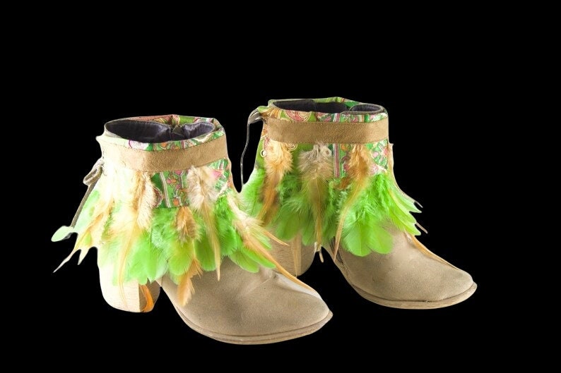 May include: A pair of beige suede ankle boots with green and brown feather trim. The boots have a block heel and a pointed toe.