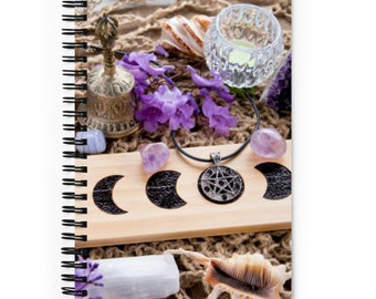 May include: A wooden board with three crescent moons painted on it. A silver necklace with a pentagram pendant hangs over the board. The board is surrounded by purple flowers, crystals, and seashells.