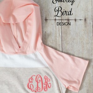 May include: A striped hooded shirt with a peach colored hood and sleeves. The shirt has a beige and white striped pattern with a peach colored monogram in the center.