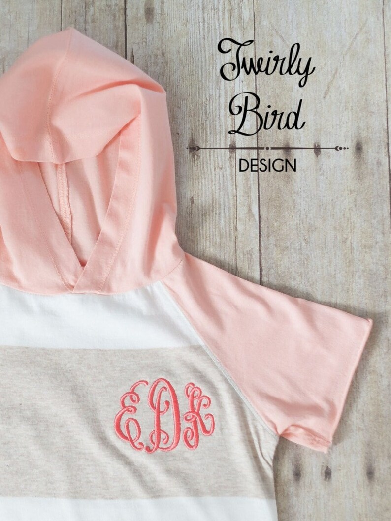 May include: A striped hooded shirt with a peach colored hood and sleeves. The shirt has a beige and white striped pattern with a peach colored monogram in the center.