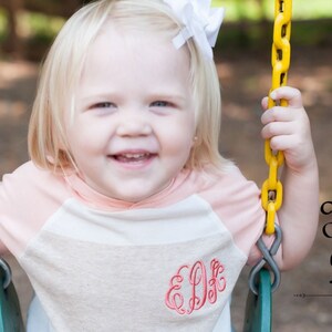 May include: A young child wearing a striped shirt with a pink and white color scheme. The shirt has a monogram embroidered on the front. The child is sitting on a swing with a yellow chain.