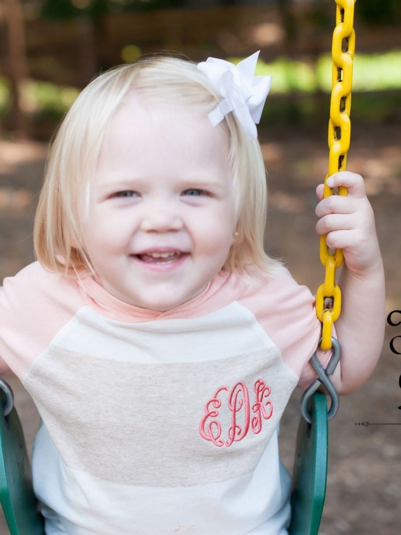 May include: A young child wearing a striped shirt with a pink and white color scheme. The shirt has a monogram embroidered on the front. The child is sitting on a swing with a yellow chain.