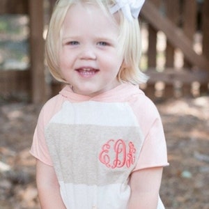 May include: A young girl with blonde hair wearing a striped, pink and white, hooded shirt with a pink monogram "E.D.K." on the front. She is wearing a white bow in her hair.