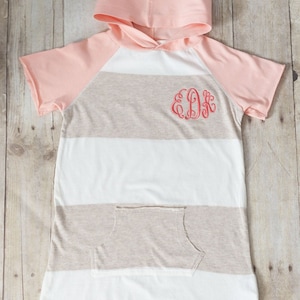 May include: A pink and beige striped hooded shirt with short sleeves and a pocket. The shirt has a monogram "EDK" embroidered on the front.