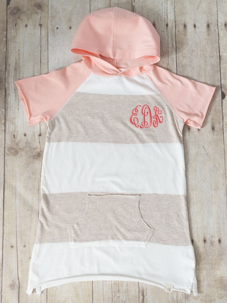May include: A pink and beige striped hooded shirt with short sleeves and a pocket. The shirt has a monogram "EDK" embroidered on the front.