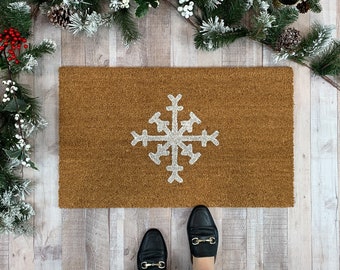 May include: A rectangular brown coir doormat featuring a centered white snowflake design. The mat is positioned on a light-colored wooden floor, surrounded by a decorative garland of green pine needles, white berries, and red holly berries.