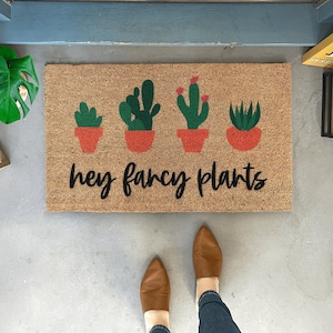 May include: A brown coir doormat with the text "hey fancy plants" in black script.  Five potted plants are depicted in green and red on the mat.  The plants are in orange pots.