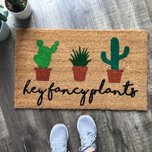 May include: A brown coir doormat with the text "hey fancy plants" in black cursive lettering. Three green cactus plants are depicted in brown pots.