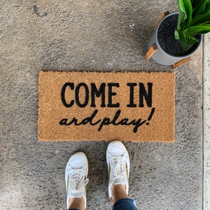 May include: A brown coir doormat with the text "COME IN and play!" in black lettering.