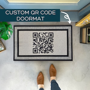 May include: A gray doormat with a black and white QR code printed on it. The doormat has a black border and is made of a textured material. The text "CUSTOM QR CODE DOORMAT" is printed in white on a blue background.