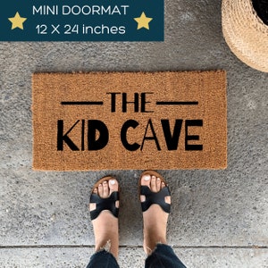 May include: A rectangular brown doormat with the text "THE KID CAVE" in black. The doormat measures 12 x 24 inches. The image also shows a person's feet wearing black sandals.
