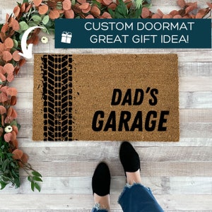 May include: A brown coir doormat with black text that reads "DAD'S GARAGE". A black tyre tread pattern is on the left side of the doormat.