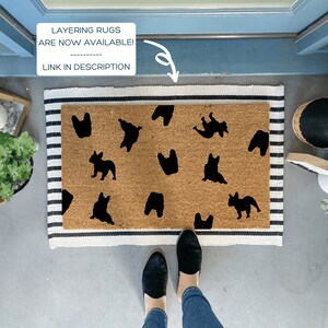 K&ouml;nnte beinhalten: Eine braune Kokosmatte mit einem schwarz-wei&szlig; gestreiften Rand. Die Fu&szlig;matte hat ein Muster aus schwarzen franz&ouml;sischen Bulldoggen-Silhouetten. Der Text "LAYERING RUGS ARE NOW AVAILABLE! LINK IN DESCRIPTION" ist auf einem wei&szlig;en Rechteck &uuml;ber der Fu&szlig;matte geschrieben.