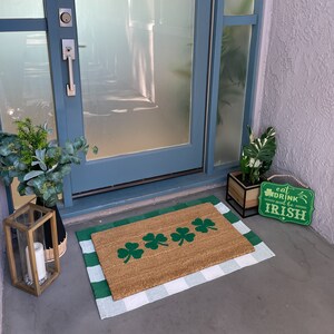 May include: A brown doormat with a white and green checkered border. The doormat has four green shamrock designs on it. The doormat is on a concrete floor in front of a glass door. A sign that says "Eat, Drink and be Irish" is on the floor next to the doormat.