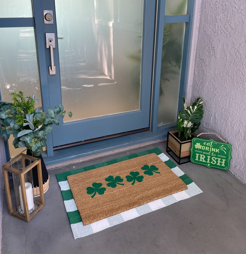 May include: A brown doormat with a white and green checkered border. The doormat has four green shamrock designs on it. The doormat is on a concrete floor in front of a glass door. A sign that says "Eat, Drink and be Irish" is on the floor next to the doormat.