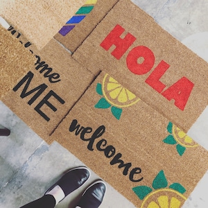 K&ouml;nnte beinhalten: Mehrere rechteckige Kokos-Fu&szlig;matten mit verschiedenen Designs. Eine Matte zeigt "WELCOME" in schwarzer Schrift mit Zitronenscheiben-Grafiken. Eine andere zeigt "HOLA" in roten Blockbuchstaben. Eine dritte zeigt "ME" in schwarzen Blockbuchstaben.