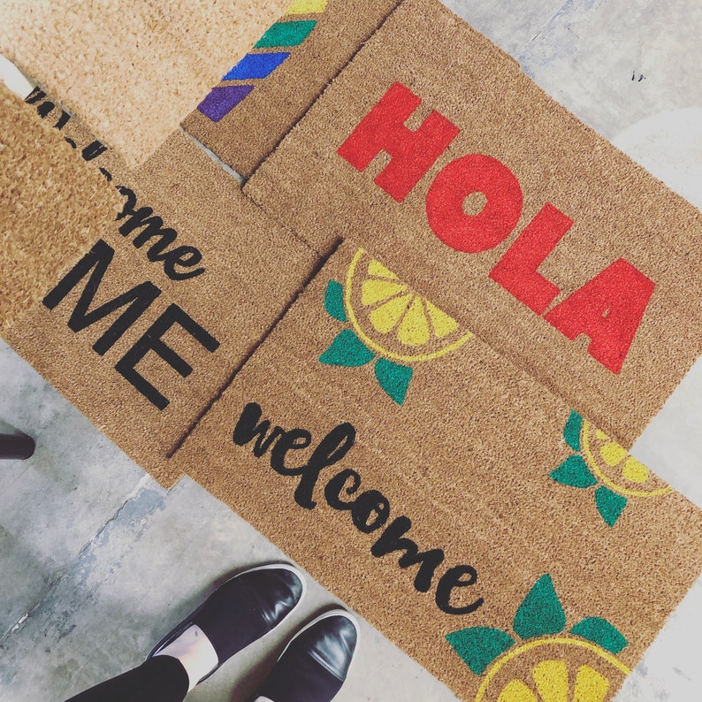 K&ouml;nnte beinhalten: Mehrere rechteckige Kokos-Fu&szlig;matten mit verschiedenen Designs. Eine Matte zeigt "WELCOME" in schwarzer Schrift mit Zitronenscheiben-Grafiken. Eine andere zeigt "HOLA" in roten Blockbuchstaben. Eine dritte zeigt "ME" in schwarzen Blockbuchstaben.