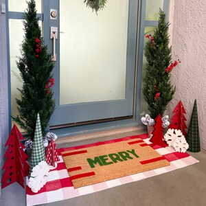 May include: A welcome mat with the word "Merry" in green letters on a brown background. The mat is placed in front of a door with two potted evergreen trees and red and white Christmas decorations.