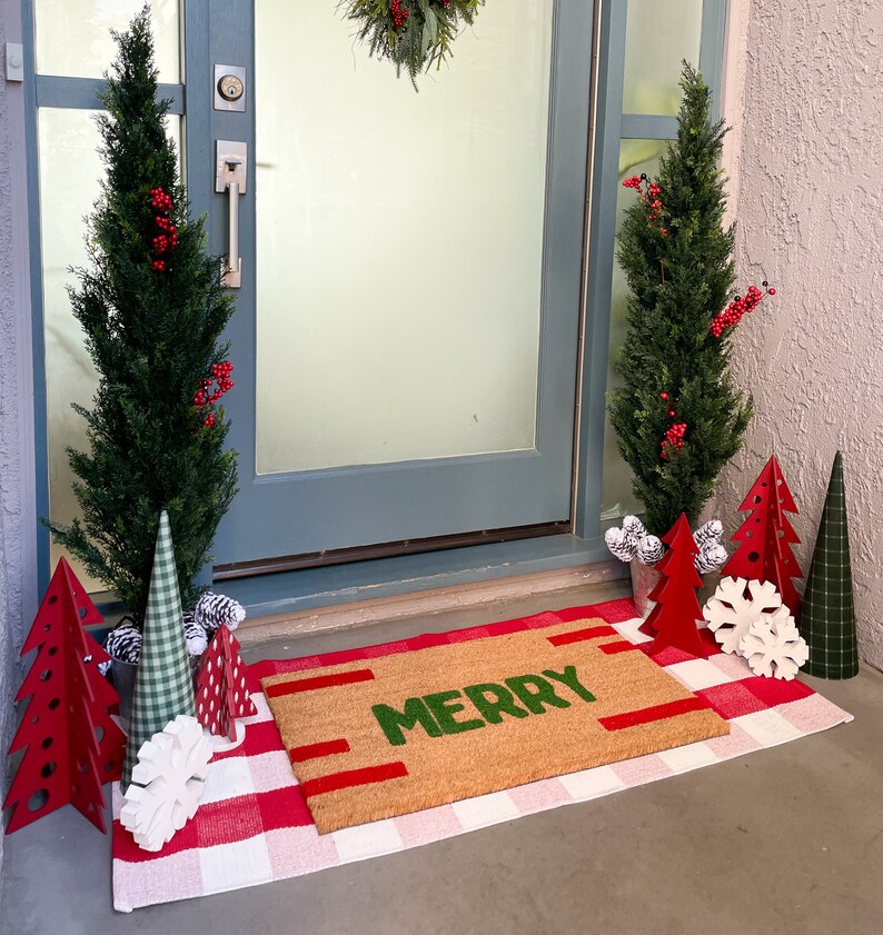 May include: A welcome mat with the word "Merry" in green letters on a brown background. The mat is placed in front of a door with two potted evergreen trees and red and white Christmas decorations.