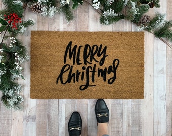 May include: A brown coir doormat with the text "Merry Christmas" in black script lettering. The doormat is on a light wood floor with green and white holiday greenery on either side.