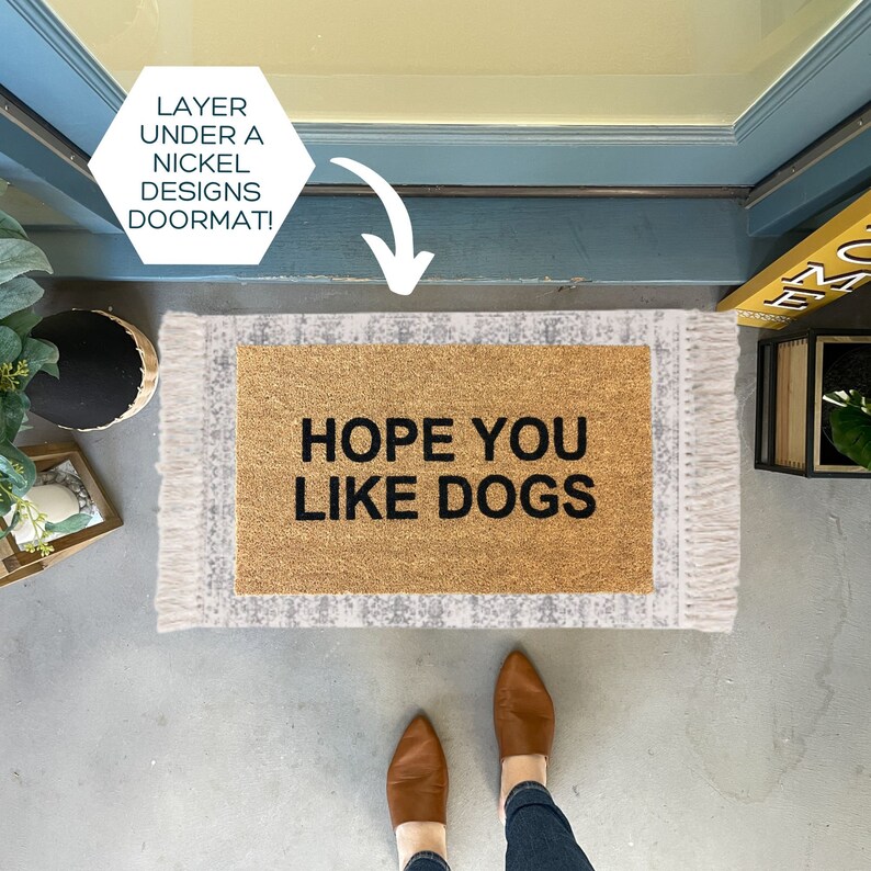 May include: A brown doormat with white fringe and the text "HOPE YOU LIKE DOGS" in black lettering. The doormat is on a gray concrete surface.