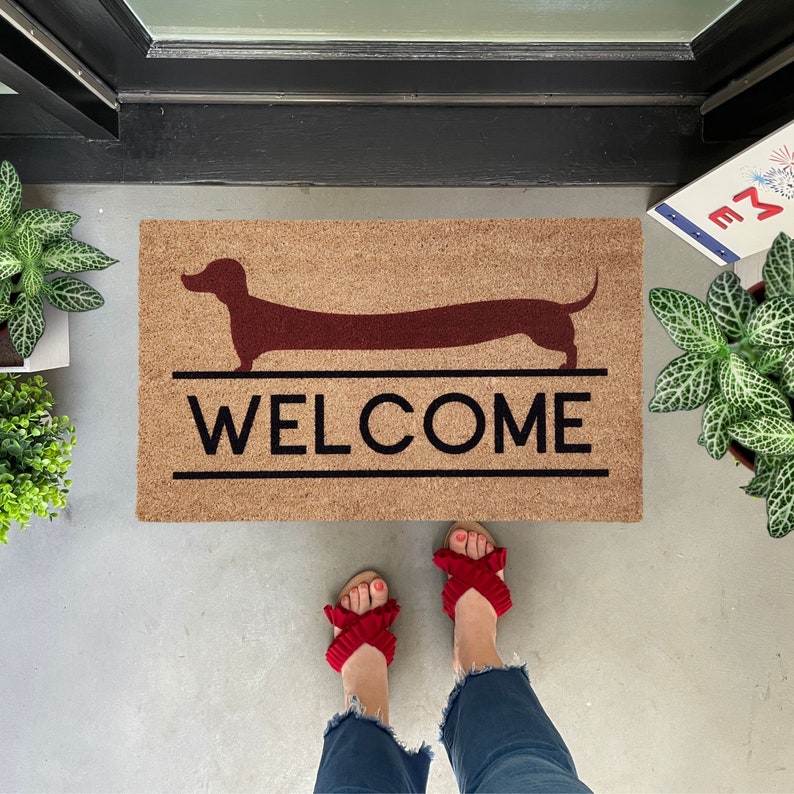 May include: A brown doormat with the word "WELCOME" printed in black letters. A silhouette of a dachshund is above the word "WELCOME". A pair of red sandals with bows are on the floor in front of the doormat.