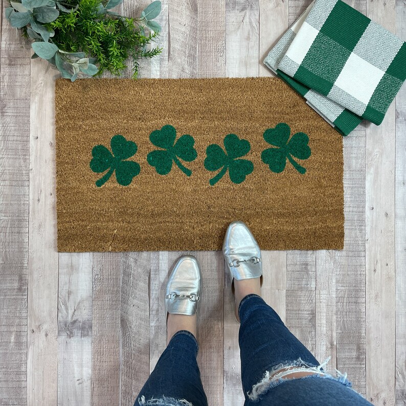 May include: A brown doormat with four green shamrock designs. The doormat is on a light wood floor. A pair of silver shoes are on the doormat.
