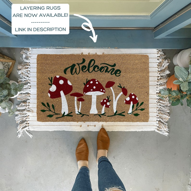 May include: A welcome mat with a brown coir surface and white fringe. The mat features a green and red mushroom design with the word "welcome" in white script.