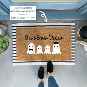 May include: A brown coir doormat with white and black stripes. The doormat has the text "Our Boo Crew" and four white ghost figures with black eyes and mouths. The ghost on the right has a black bow.
