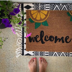 K&ouml;nnte beinhalten: Eine braune Fu&szlig;matte mit einem Zitrusfrucht-Design und dem Wort "welcome" in schwarzer Schrift. Die Matte liegt auf einem gemusterten Teppich mit einem schwarz-wei&szlig;en geometrischen Design. Violette Blumen sind in der oberen linken Ecke zu sehen.