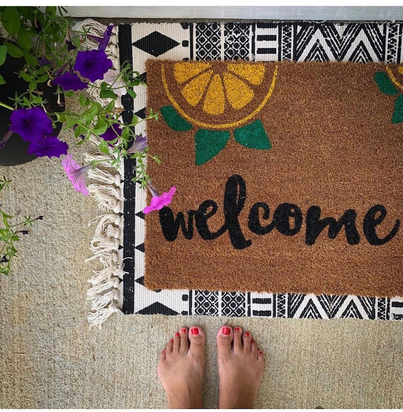 K&ouml;nnte beinhalten: Eine braune Fu&szlig;matte mit einem Zitrusfrucht-Design und dem Wort "welcome" in schwarzer Schrift. Die Matte liegt auf einem gemusterten Teppich mit einem schwarz-wei&szlig;en geometrischen Design. Violette Blumen sind in der oberen linken Ecke zu sehen.