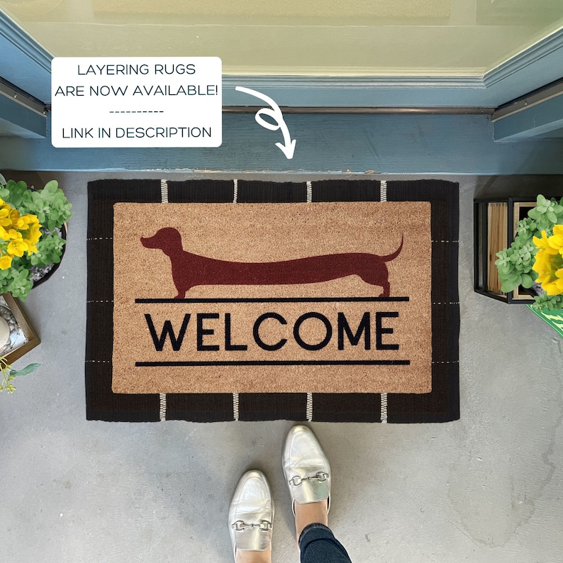 May include: A brown coir welcome mat with black trim features a red dachshund silhouette and the word "WELCOME" in black lettering.