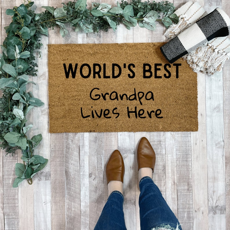 May include: A brown coir doormat with the text "WORLD'S BEST Grandpa Lives Here" in black lettering.