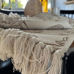 May include: A cream-colored woven table runner featuring a brown botanical line art pattern and long, knotted fringe trim along the edges. The fabric has a visible textured weave, and the design includes repeating floral and leaf motifs.