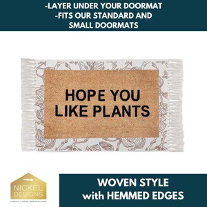 May include: A rectangular doormat layer featuring a central tan coir section with the black text HOPE YOU LIKE PLANTS. The border is white fabric with a brown floral line drawing pattern and fringed edges on the left and right sides.