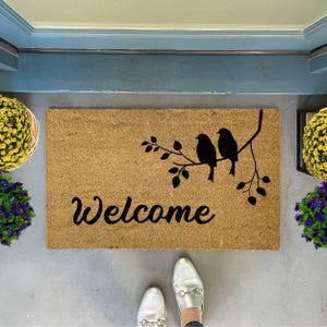 Puede incluir: Una alfombra de coco marr&oacute;n con la palabra "Welcome" en letras cursivas negras. Dos p&aacute;jaros negros est&aacute;n posados en una rama con hojas, tambi&eacute;n en negro, sobre la palabra "Welcome".