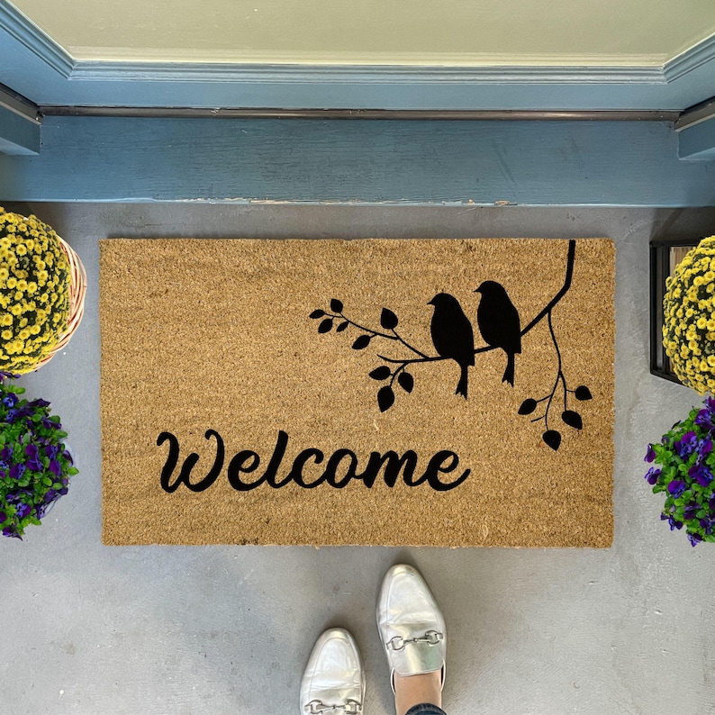 Puede incluir: Una alfombra de coco marr&oacute;n con la palabra "Welcome" en letras cursivas negras. Dos p&aacute;jaros negros est&aacute;n posados en una rama con hojas, tambi&eacute;n en negro, sobre la palabra "Welcome".