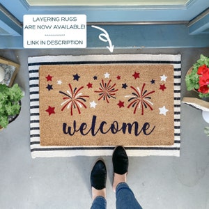 May include: A welcome mat with a red, white, and blue firework design and the word "welcome" in black script. The mat has a striped border in black and white.