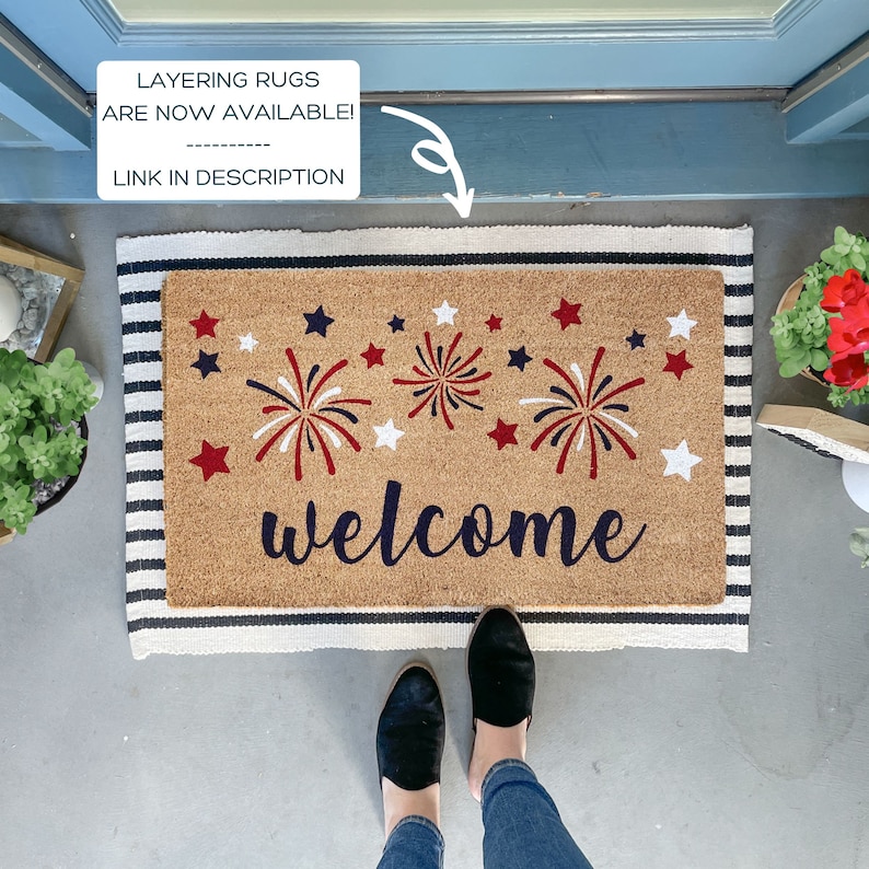 May include: A welcome mat with a red, white, and blue firework design and the word "welcome" in black script. The mat has a striped border in black and white.