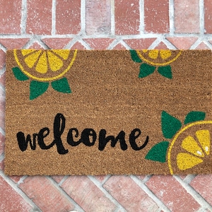K&ouml;nnte beinhalten: Rechteckige Fu&szlig;matte aus braunem Kokosfasermaterial. Der Schriftzug "welcome" ist in schwarzer Schrift in der Mitte zu sehen. Die Matte ist mit gelben und gr&uuml;nen Zitronenscheiben-Designs in jeder Ecke verziert.